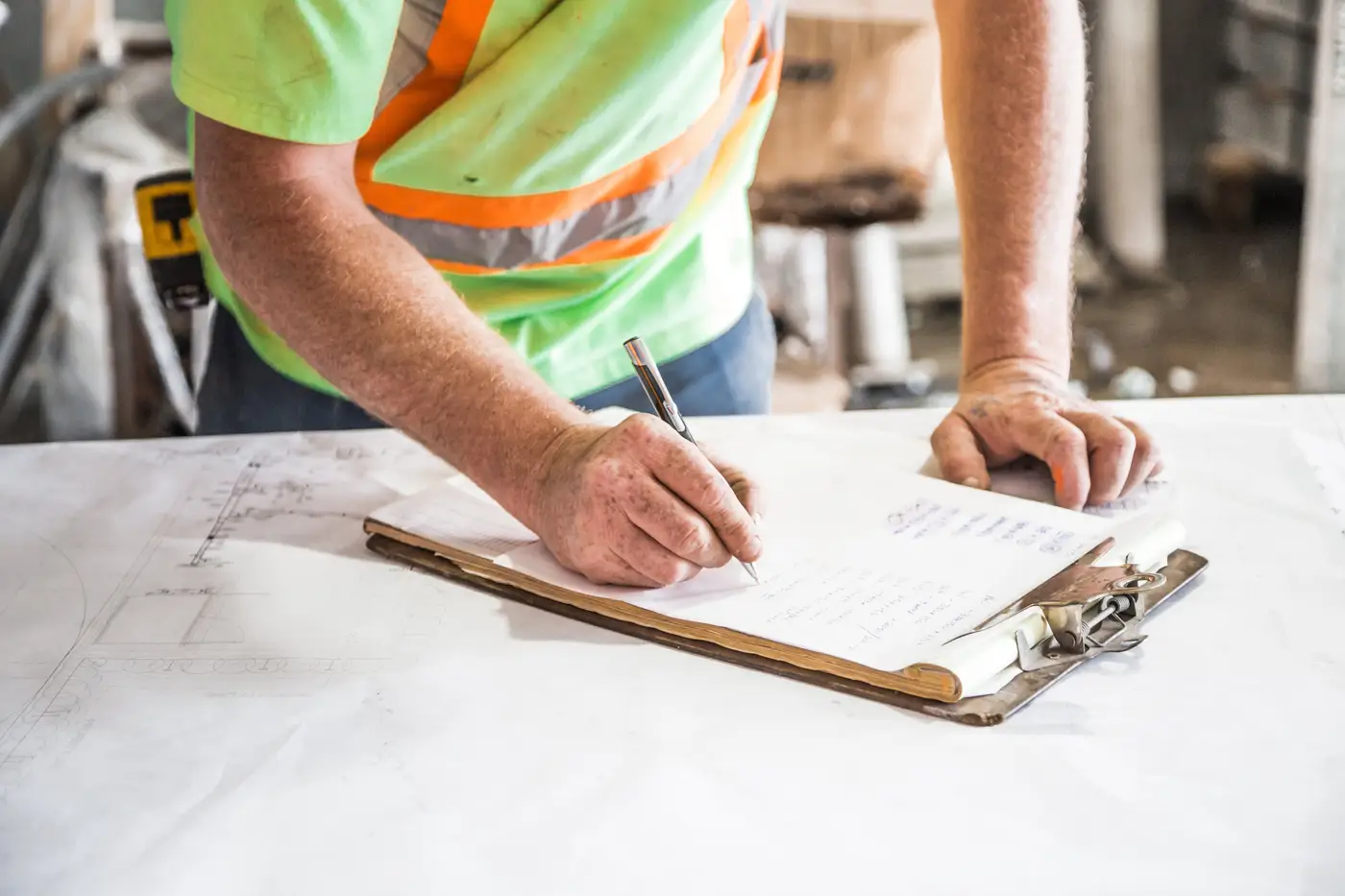A worker in a safety vest writes notes on a clipboard beside rolled site plans on a jobsite table.