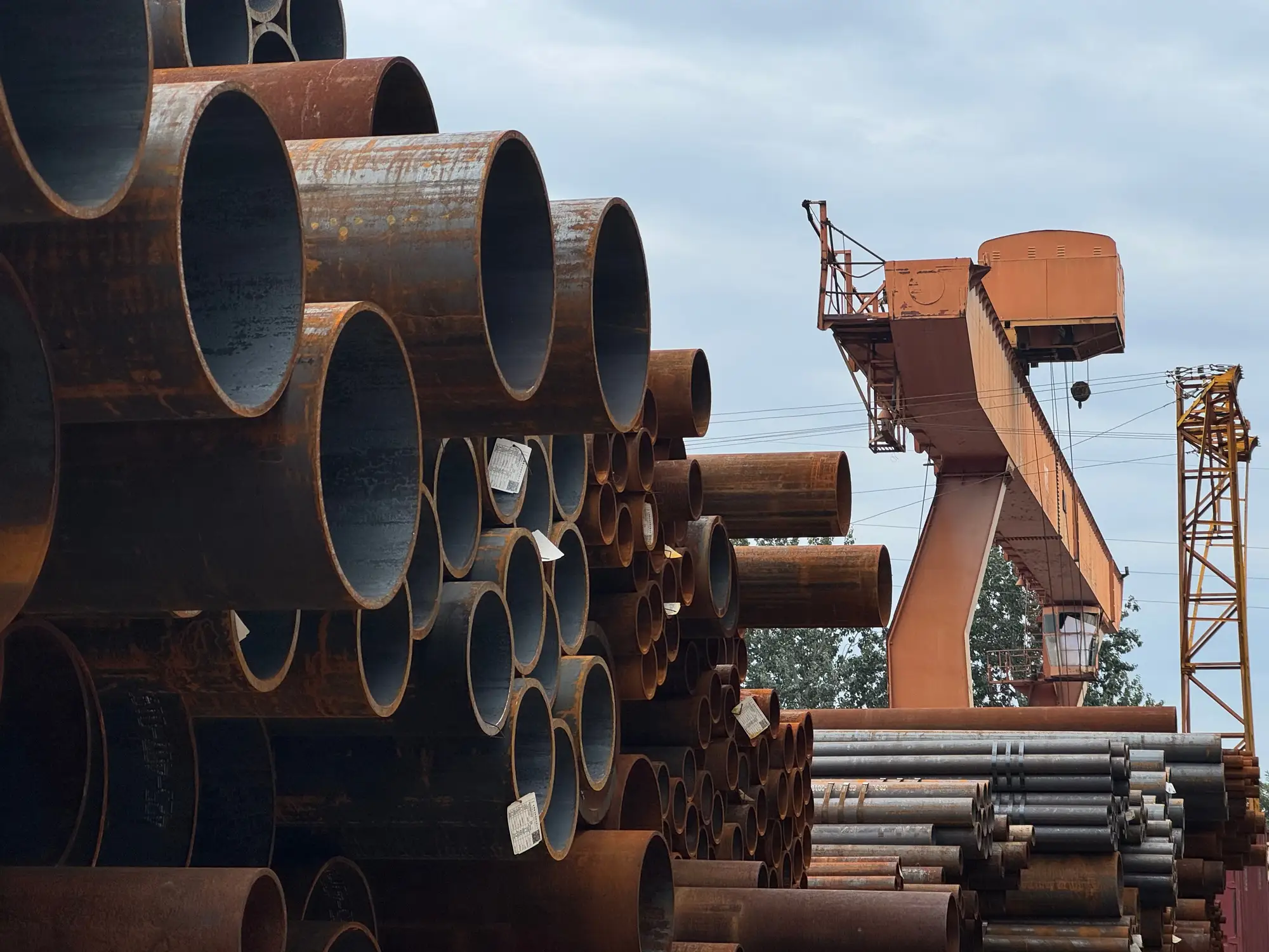 Stacked lengths of rusted steel pipe in an outdoor yard beside a gantry crane