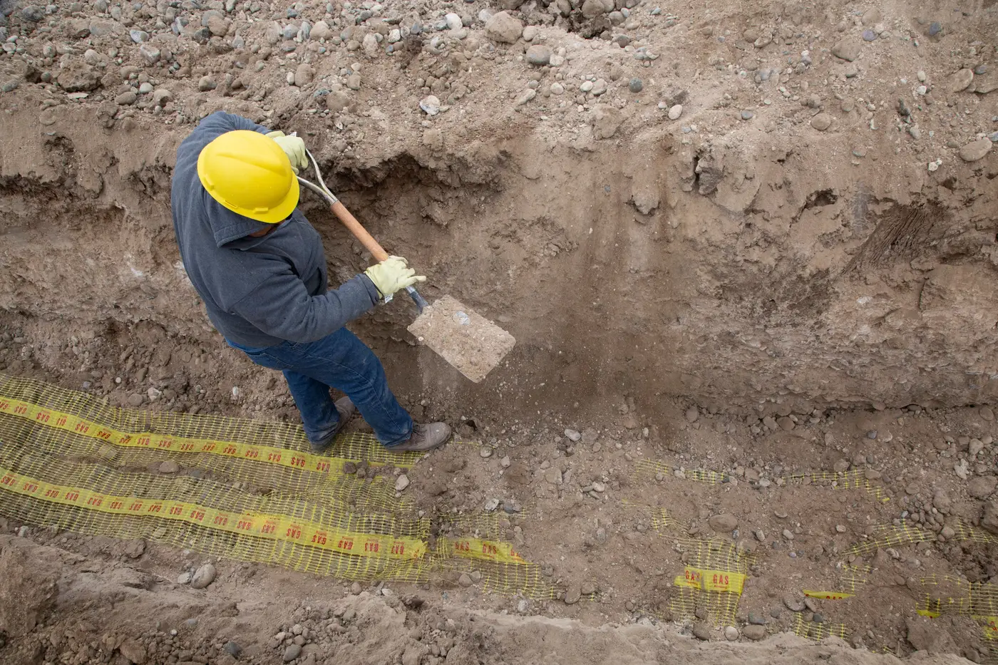 Worker digging by hand in a narrow trench, seen from above, with yellow caution tape along the edge.